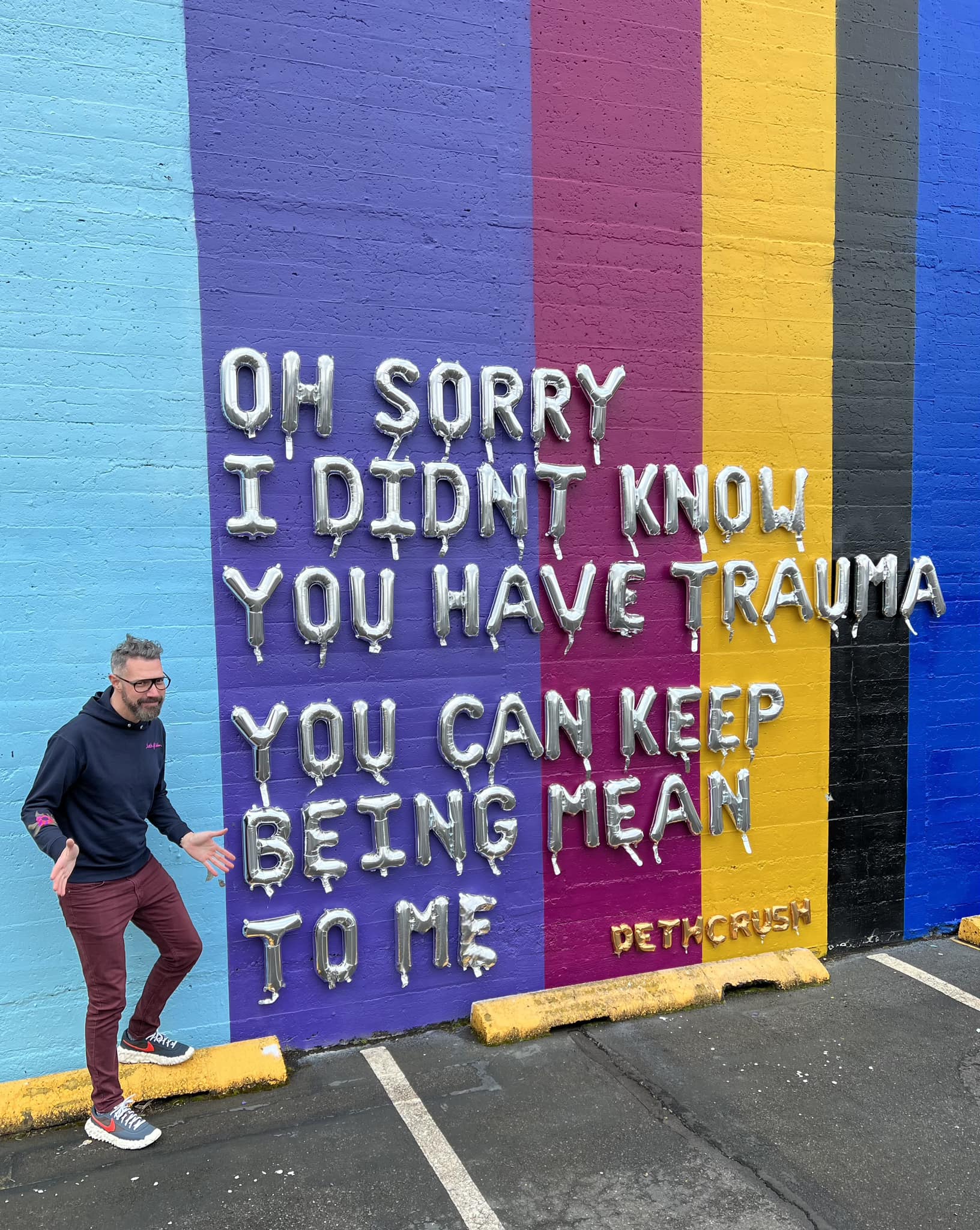 The meme is a photo of a man standing in front of a colorful wall with a quote written in large, dripping silver letters. The quote reads: "OH SORRY I DIDNT KNOW YOU HAD TRAUMA YOU CAN KEEP BEING MEAN TO ME". The image is humorous and ironic, implying that the person is not interested in engaging with someone who is being mean or hurtful.
